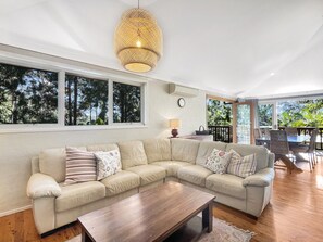 Un salon doté d’un grand canapé en forme de L, d’une table basse en bois et d’un luminaire suspendu. De grandes fenêtres offrent une vue sur les arbres, et un balcon aménagé avec du mobilier d’extérieur complète l’ensemble.