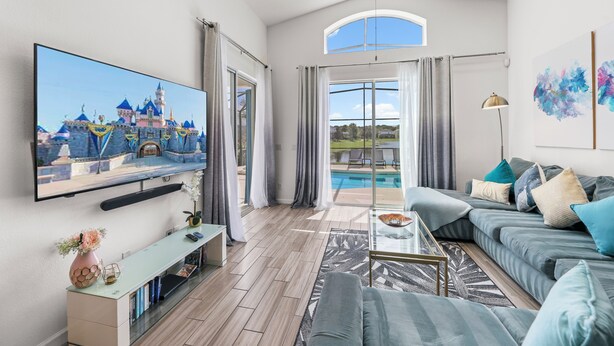 A modern living room with a large flat-screen TV, a grey sofa, a coffee table, and a view of a swimming pool. - 4BR Retreat, Pool, Close to Disney (Kissimmee)