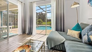 A spacious living room with a large glass door leading to an outdoor area, a cozy sofa with cushions, and a modern floor lamp. - 4BR Retreat, Pool, Close to Disney (Kissimmee)