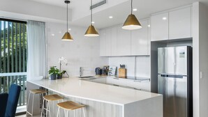 A modern kitchen with a large island, stainless steel appliances, pendant lighting, and a view of greenery outside. - Spacious and Superb 3-bed Apartment (Brisbane)