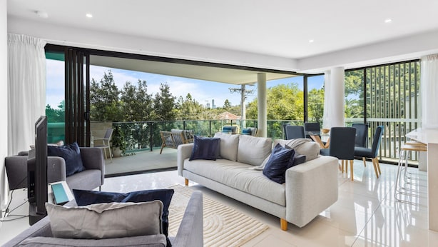 A modern living room with a sofa, chairs, and a flat-screen TV. There is a balcony with outdoor furniture and a view of trees and buildings. - Spacious and Superb 3-bed Apartment (Brisbane)