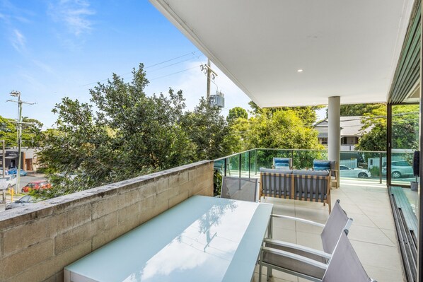 A balcony with a glass railing, a seating area with a table and chairs, and a view of trees and buildings. - Spacious and Superb 3-bed Apartment (Brisbane)