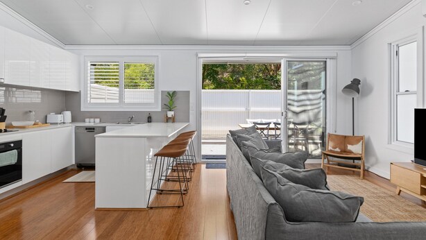 A modern kitchen with white cabinets, a central island with bar stools, and a large sliding glass door leading to a patio with outdoor furniture. - Relaxing 2-bed Home Close to Beaches and Parks (Sydney)