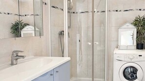 Ett modernt badrum med en dusch som är avskärmad med glasväggar, ett handfat med blandare, en tvättmaskin och en krukväxt.