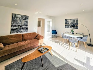 A living room with a brown sofa, a coffee table, a dining area with a round table and chairs, and two black-and-white paintings on the walls.