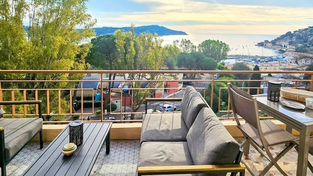 En takterrass med en soffa, ett matbord och stolar, med utsikt över ett kustnära område med byggnader och träd.