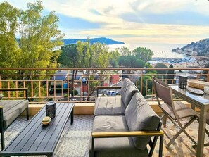 Terraza en la azotea con sofá, mesa de comedor y sillas, con vistas a una zona costera con edificios y árboles.