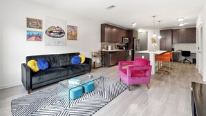 Una sala de estar moderna con un sofá negro, cojines coloridos, una mesa de centro de vidrio y un tapete geométrico.