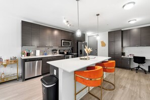 Une cuisine moderne avec des meubles en bois foncé, des appareils électroménagers en acier inoxydable et un îlot central blanc agrémenté de tabourets de bar orange.
