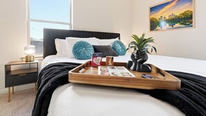 Una cama bien hecha con una bandeja de libros y un control remoto, una lámpara de noche y una planta en maceta sobre una mesita de noche.