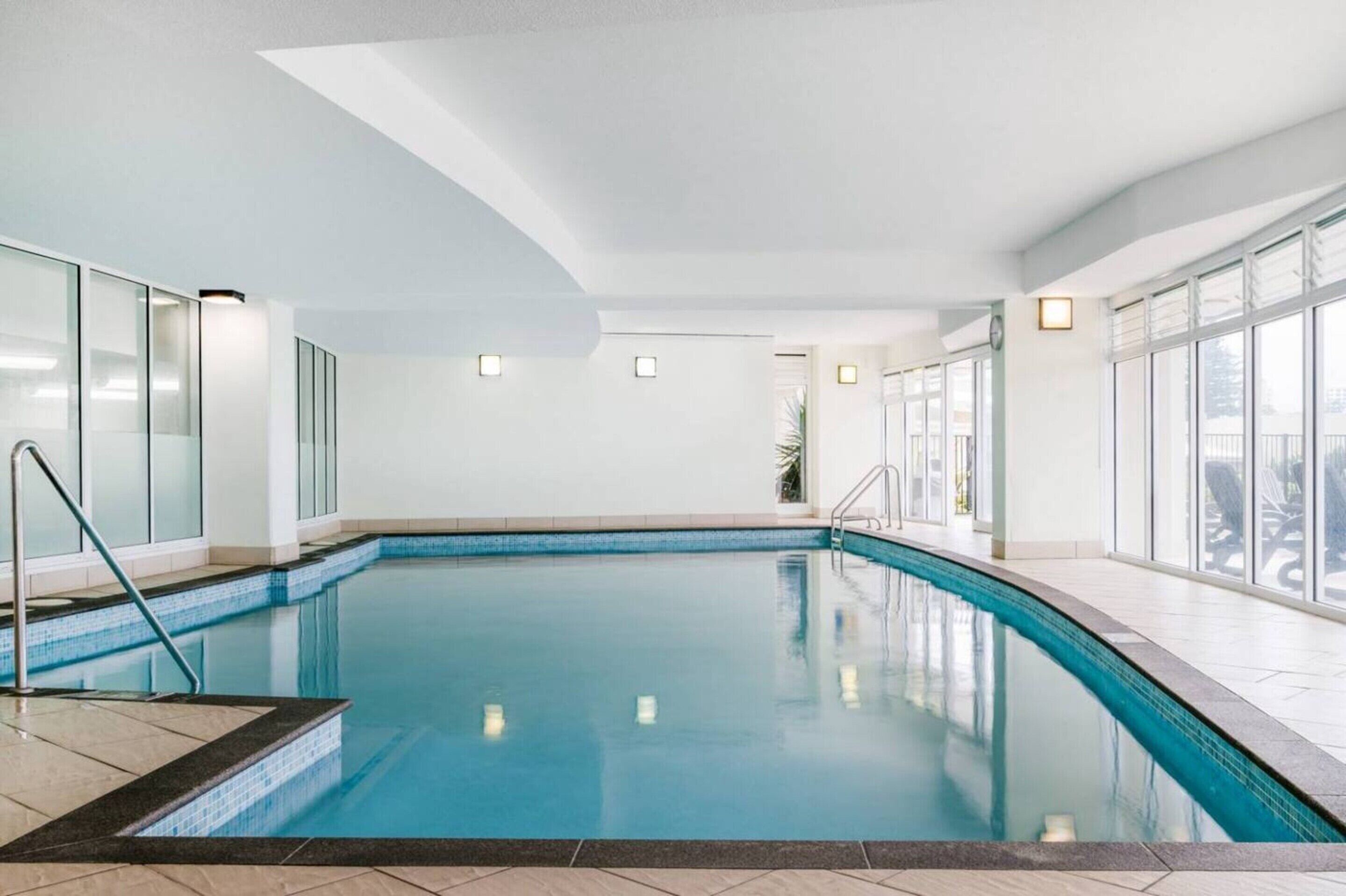 An indoor swimming pool with clear glass walls, a tiled floor, and a ceiling with recessed lighting.