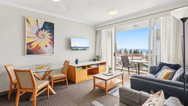 Une chambre d’hôtel moderne avec balcon, un coin repas doté d’une table en verre, un canapé et un téléviseur fixé au mur.