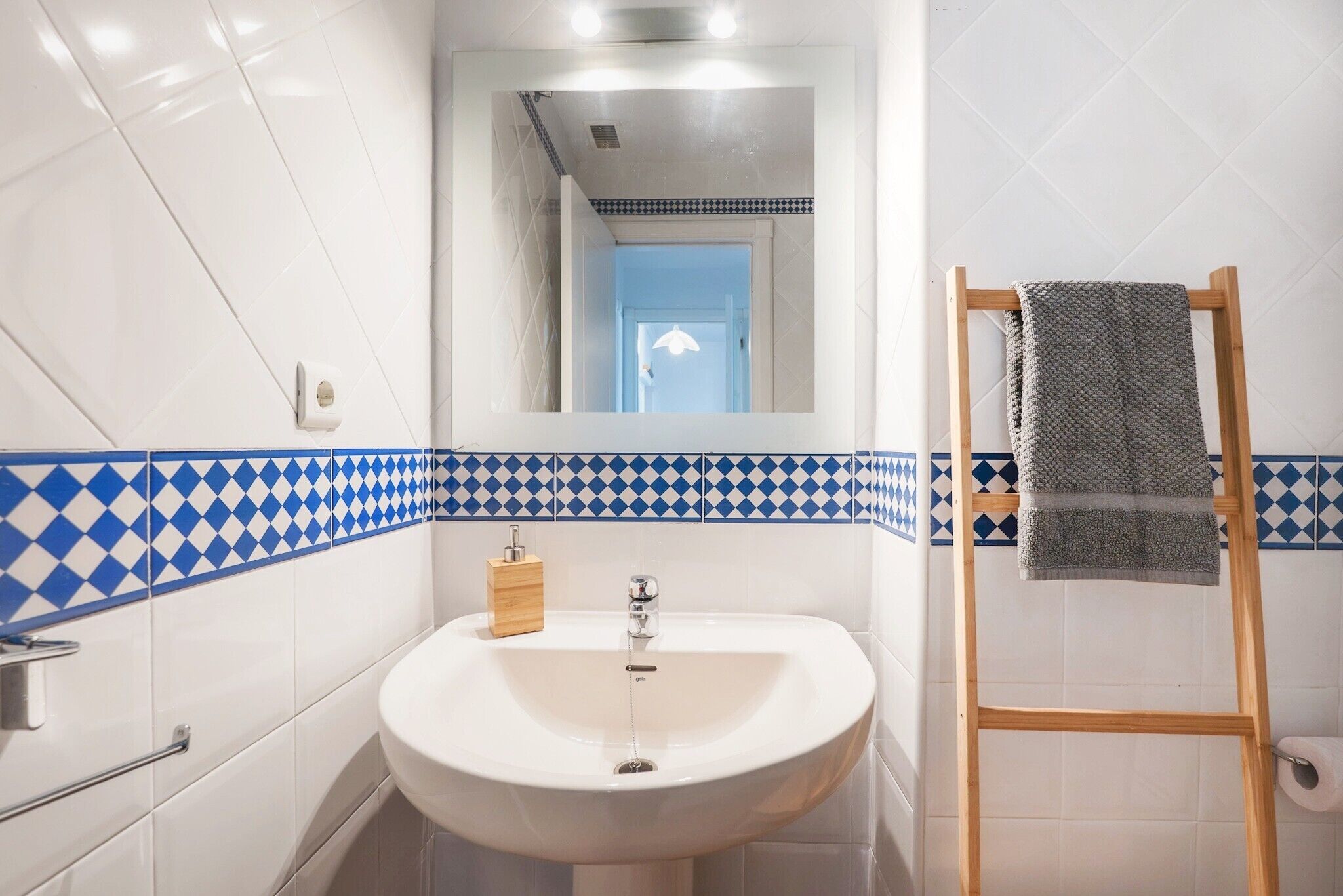 A bathroom with a white sink, a wooden soap dispenser, a towel rack with a towel, and a blue and white patterned tile border.