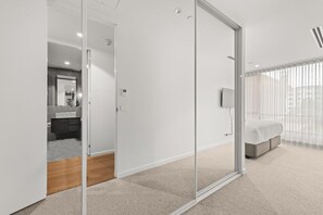 A modern bedroom with a glass door leading to a bathroom, a bed with a grey headboard, and a large window with sheer curtains.