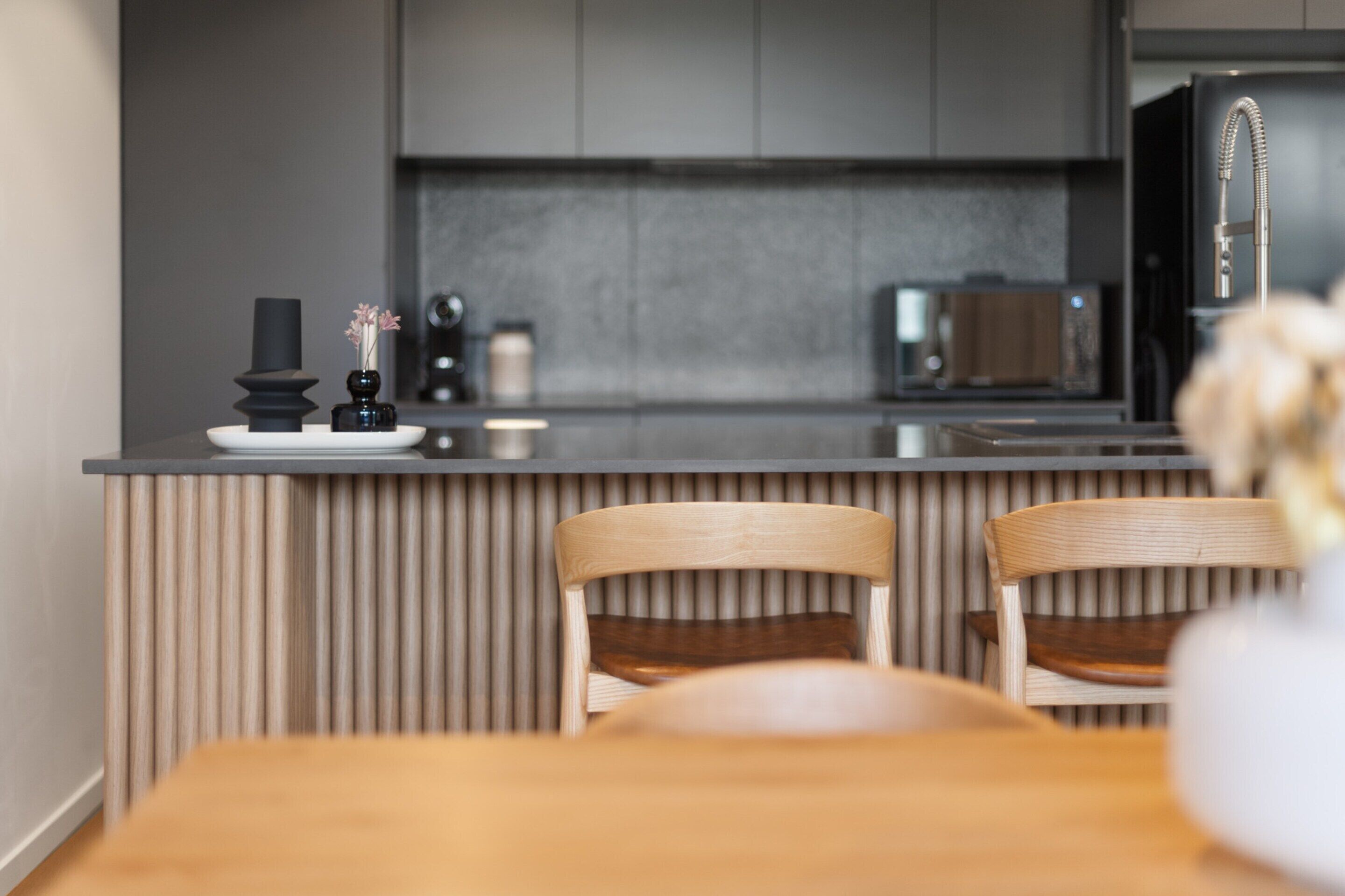 A modern kitchen with a wooden dining table and chairs, a black vase with flowers, and a microwave.