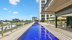 Uma piscina infinita no terraço com vista desobstruída para o horizonte da cidade, rodeada por espreguiçadeiras e guarda-sóis.