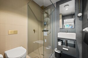 Un baño moderno con ducha de vidrio, inodoro blanco y tocador oscuro con lavamanos y espejo.