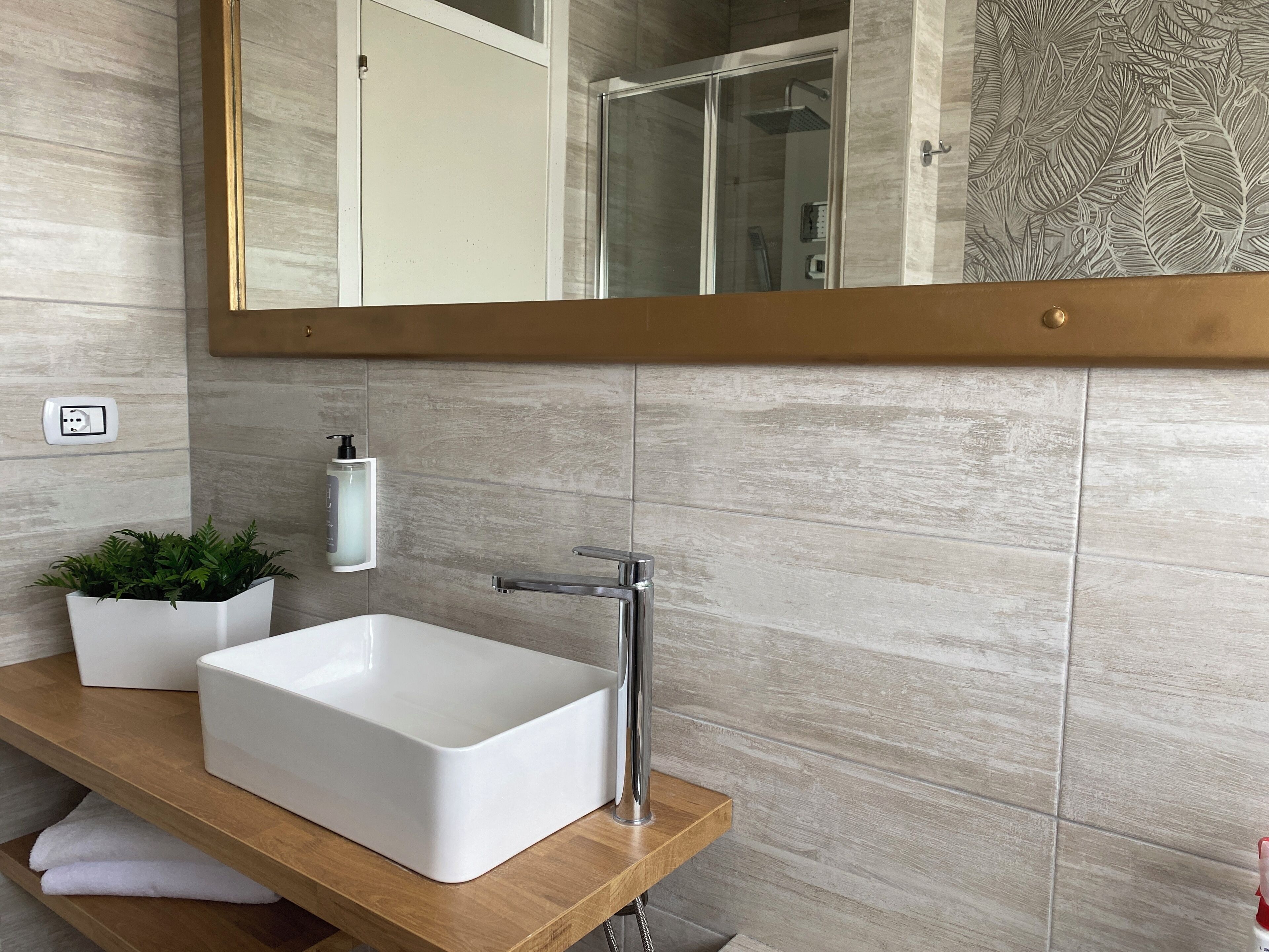 Un baño moderno con un lavamanos rectangular blanco, una llave de cromo, un mueble de tocador de madera, una planta en maceta y un espejo grande con marco dorado.