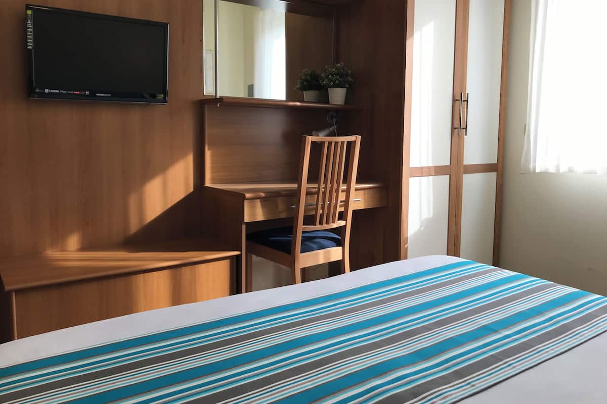 Habitació amb un llit, escriptori, cadira, televisió i finestra amb cortines.