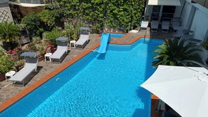En pool med solstolar och en vattenrutschkana, omgiven av grönska och en stenmur.