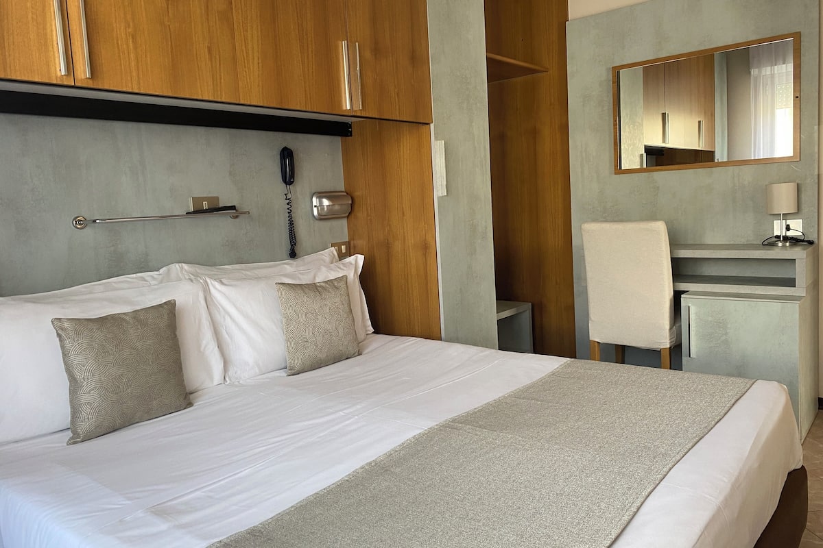 Habitació d'hotel amb un llit gran, escriptori, cadira, mirall, telèfon i penjador de tovalloles.