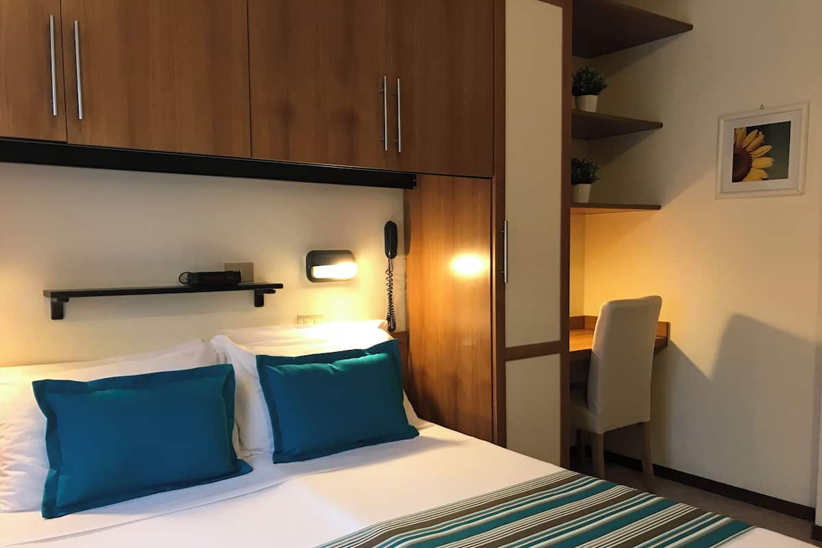 Una habitació d'hotel amb un llit, una taula de nit i una cadira. Hi ha un telèfon a la paret i una estant amb plantes.