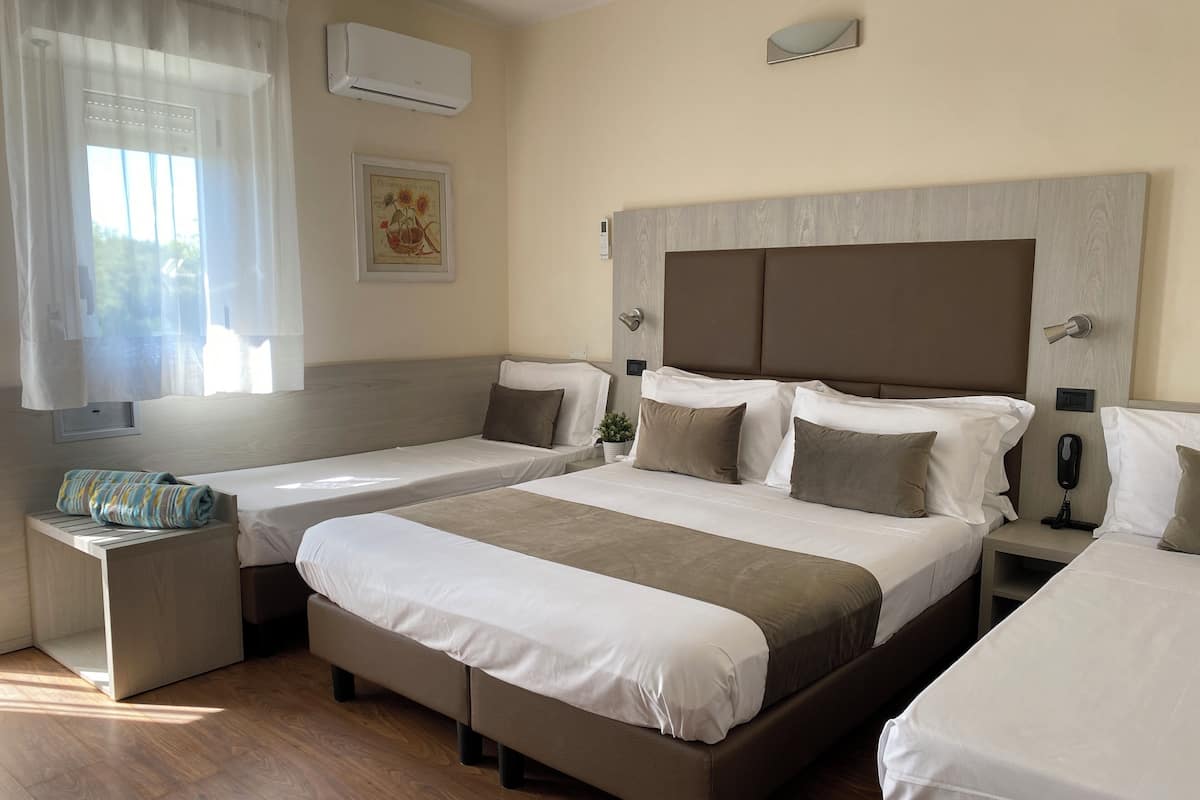 Habitació d'hotel amb dos llits individuals i un de més gran, tots amb roba de llit blanca. Hi ha una petita taula de nit amb una làmpada i un telèfon a sobre. Una finestra amb cortines de tela fina deixa entrar llum natural a l'habitació.