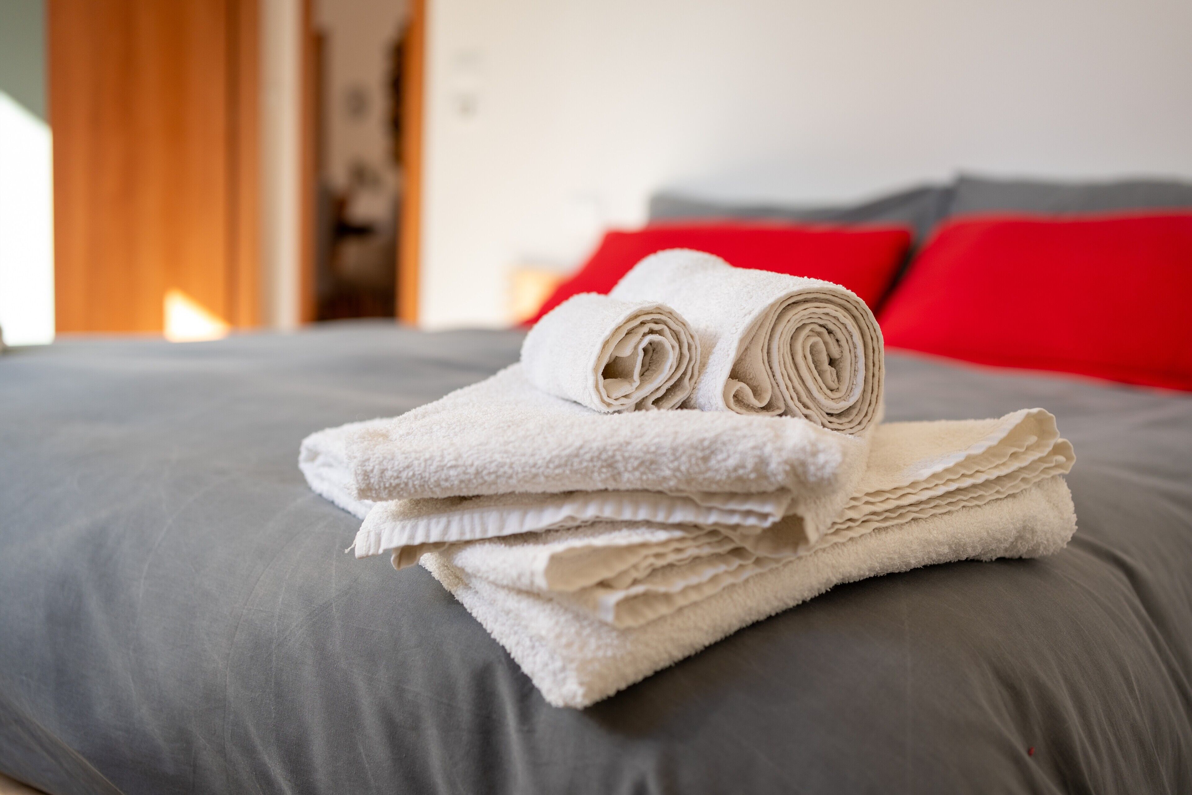 Un montón de toallas blancas dobladas ordenadamente sobre una cama gris con almohadas rojas.