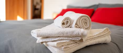 Un montón de toallas blancas dobladas ordenadamente sobre una cama gris con almohadas rojas.