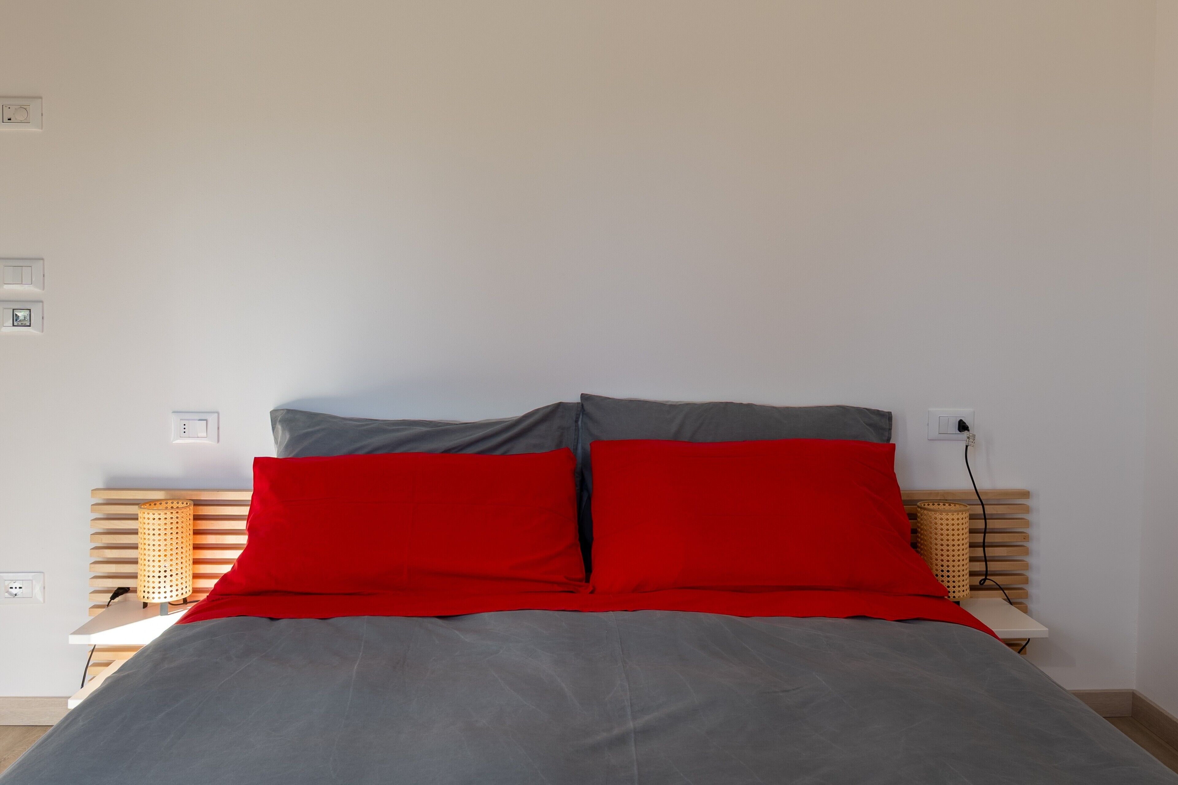 A bed with grey bedding and red pillows, a bedside lamp, and a power outlet on the wall.