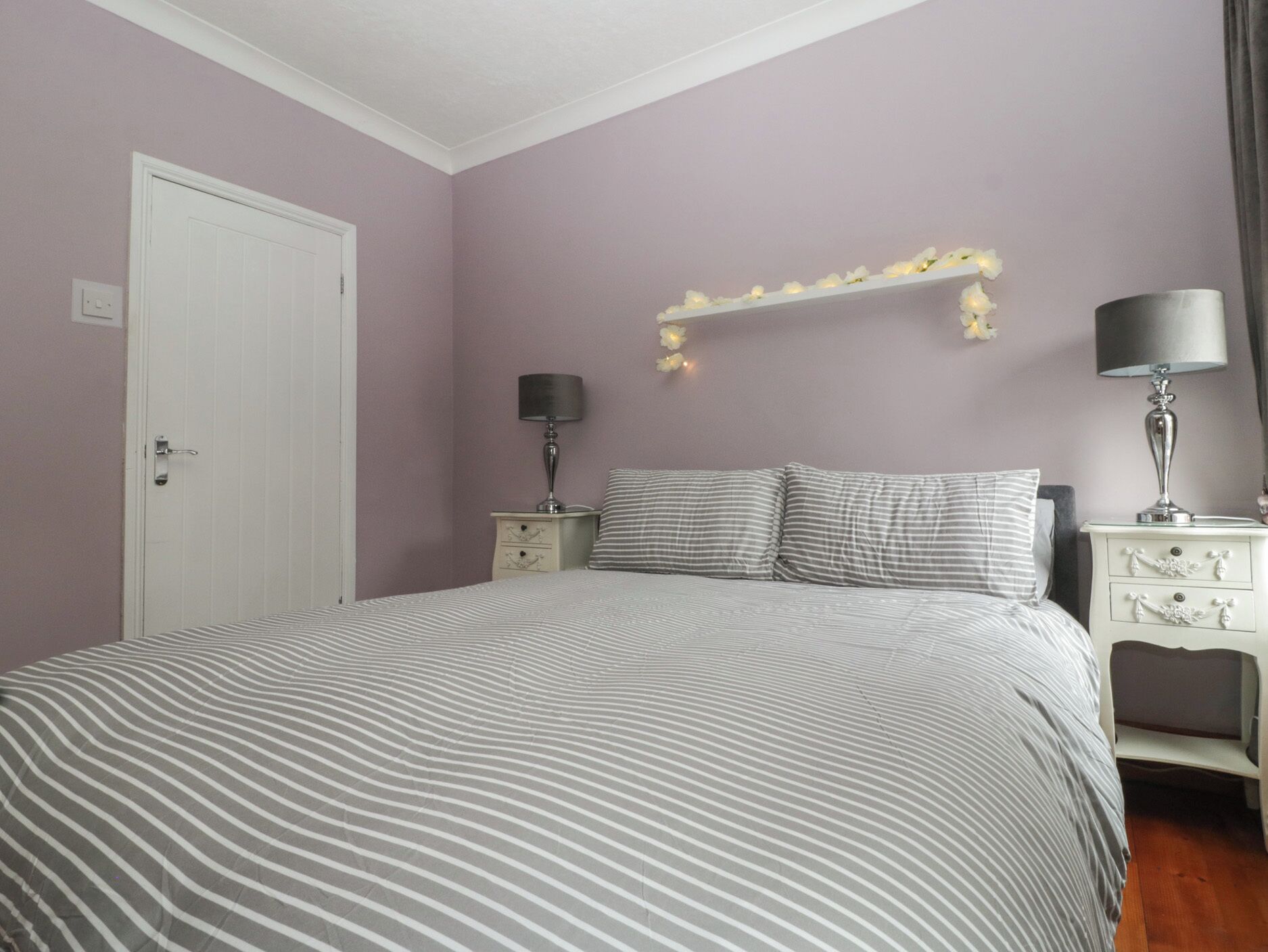 A bedroom with a bed, two bedside tables, a nightstand, a wall-mounted light fixture, and a door.