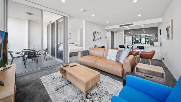 A modern living room with a sofa, coffee table, and blue armchair. There is a dining area with a table and chairs, and a kitchen in the background. - Stylish 1-bed Retreat Steps From Foreshore (Canberra)