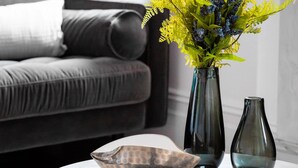 Un salon avec un canapé gris, une table ronde sur laquelle se trouvent un bol et un livre, ainsi que deux vases avec des plantes.