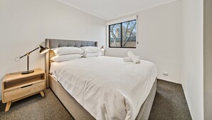 Un dormitorio moderno con una cama grande, una mesita de noche con lámpara y una ventana con vistas a edificios.