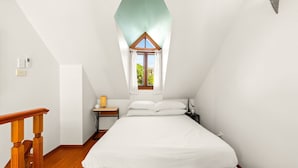 Une chambre avec un plafond en pente, une fenêtre avec des rideaux, un cadre de lit en bois, une table de chevet avec une lampe et une commode.