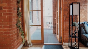 A room with a sliding glass door leading to a balcony, a brick wall, a black metal shelf with decorative items, and a blue sofa.