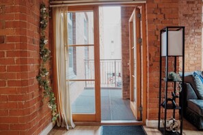 A room with a sliding glass door leading to a balcony, a brick wall, a black metal shelf with decorative items, and a blue sofa.