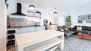 Eine moderne Küche mit weißer Arbeitsplatte, schwarzem Dunstabzug und Pendelleuchten. Im Wohnbereich befindet sich ein Esstisch mit Bänken, eine Couch und ein Couchtisch.