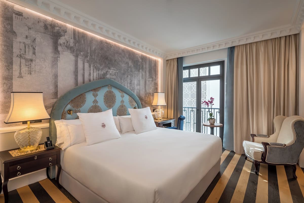 A hotel room with a large bed, two bedside tables with lamps, a chair, a view of the outdoors, and a decorative wall.