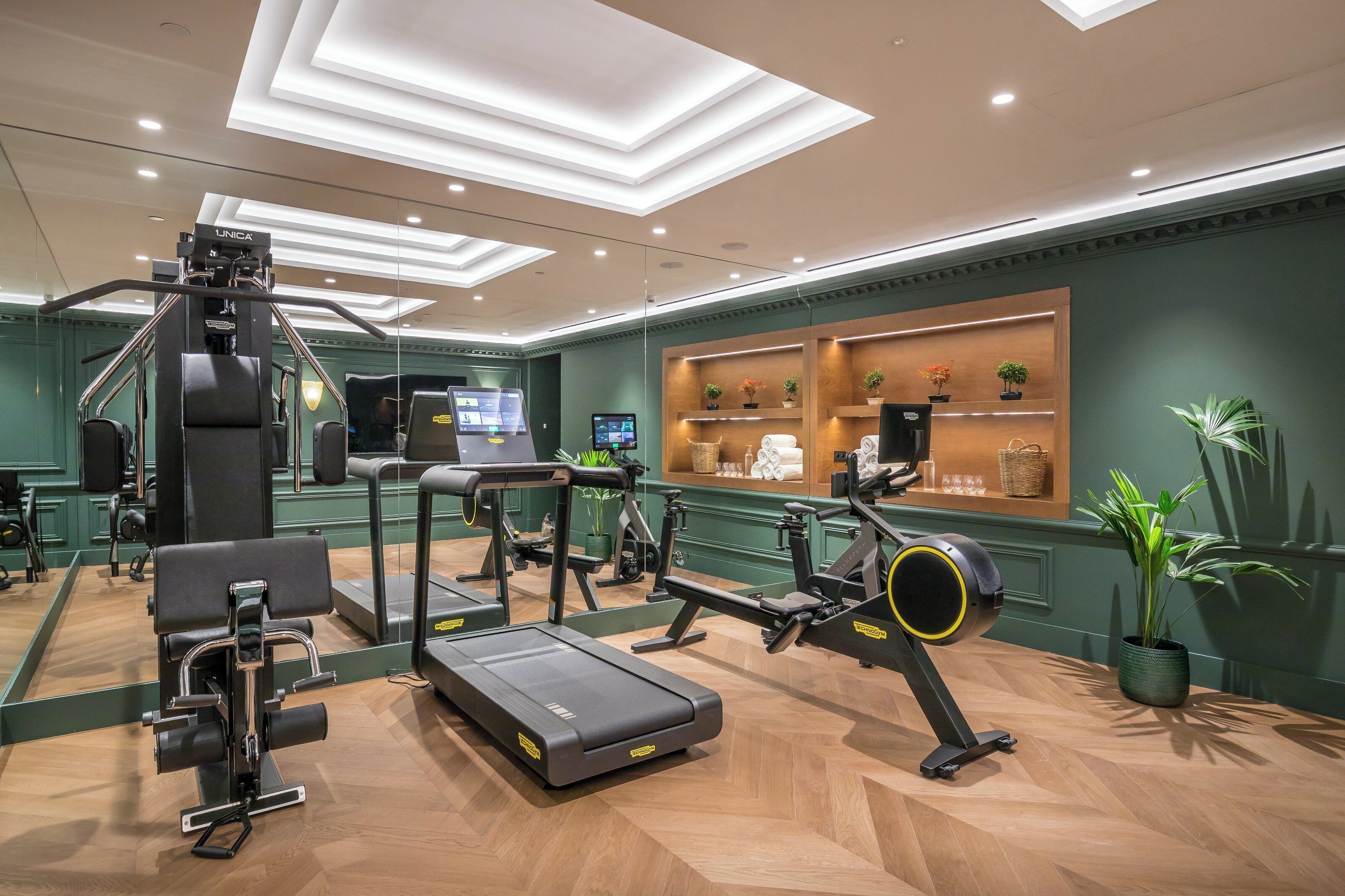 Un gimnasio con equipo de ejercicio, un quiosco digital y una zona de recepción con estanterías y plantas.