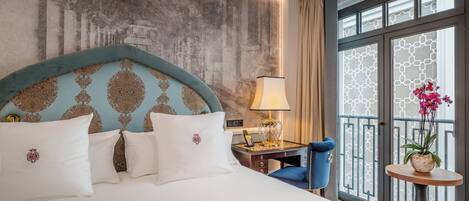 Ein Hotelzimmer mit einem großen Bett, einem Schreibtisch mit Lampe, einem Sessel, einem kleinen Tisch mit Pflanze und einem Fenster mit Blick.