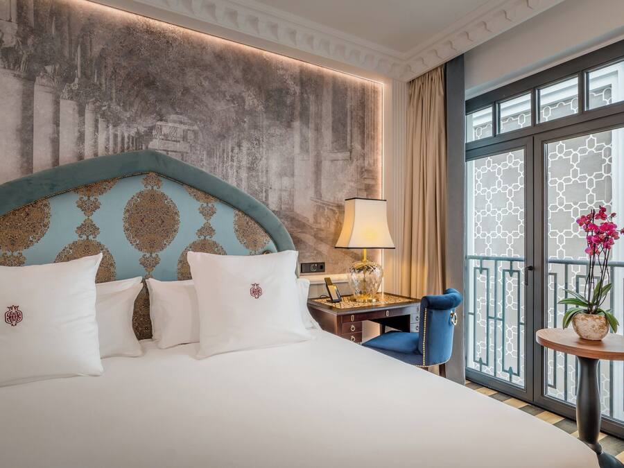 Habitación de hotel con una cama grande, un escritorio con lámpara, una silla, una mesita con una planta y una ventana con vistas.
