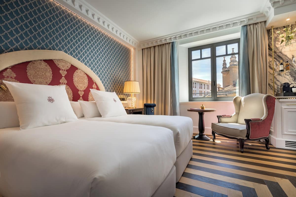 A hotel room with a large bed, a chair, a small table, and a view of a cityscape through the window.