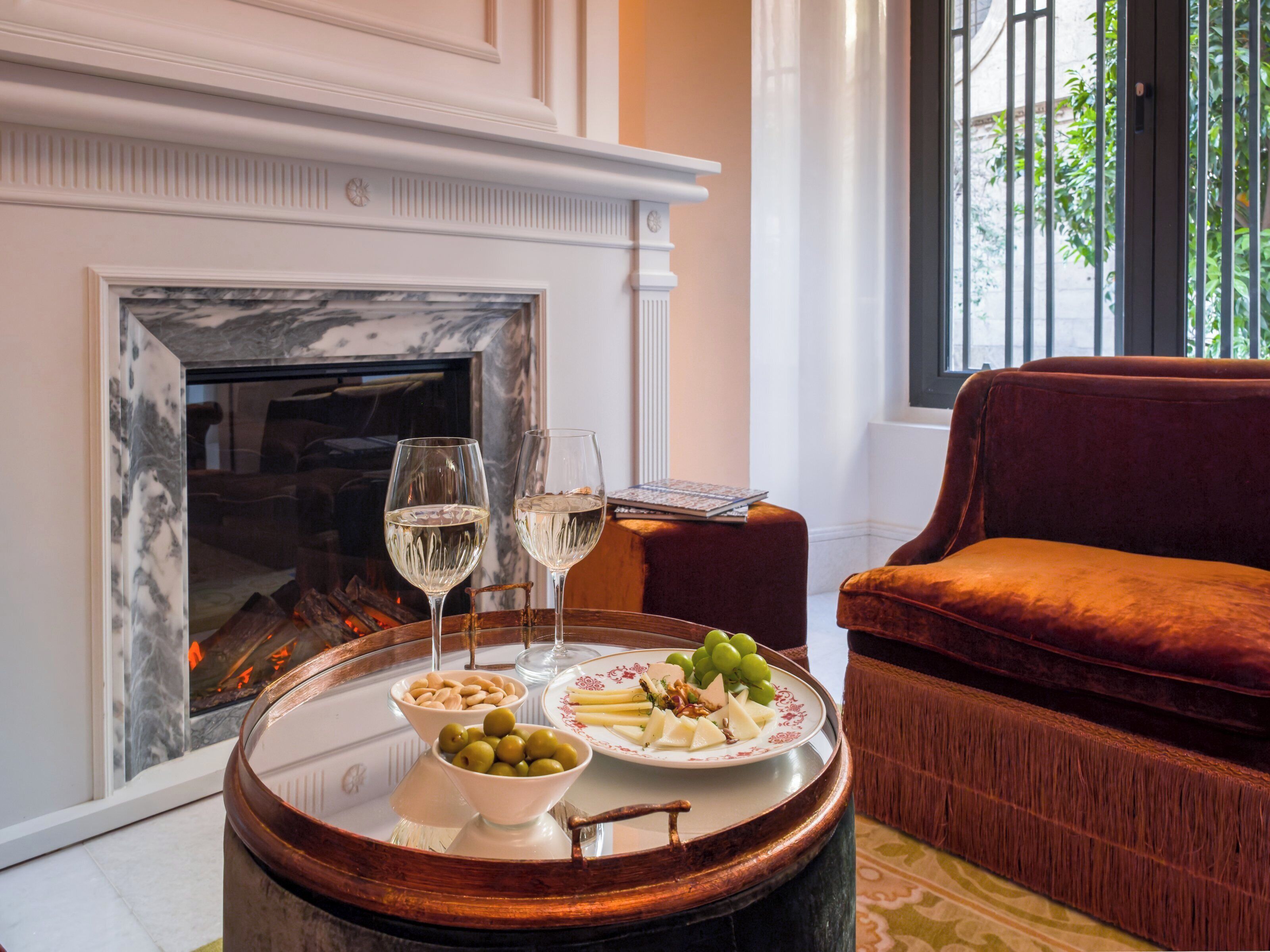 Una habitación con chimenea, una mesa redonda de madera con una bandeja de comida y dos copas de vino blanco, y un sofá de terciopelo rojo.