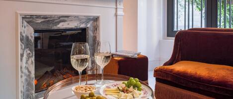 A room with a fireplace, a round wooden table with a tray of food and two glasses of white wine, and a red velvet sofa.