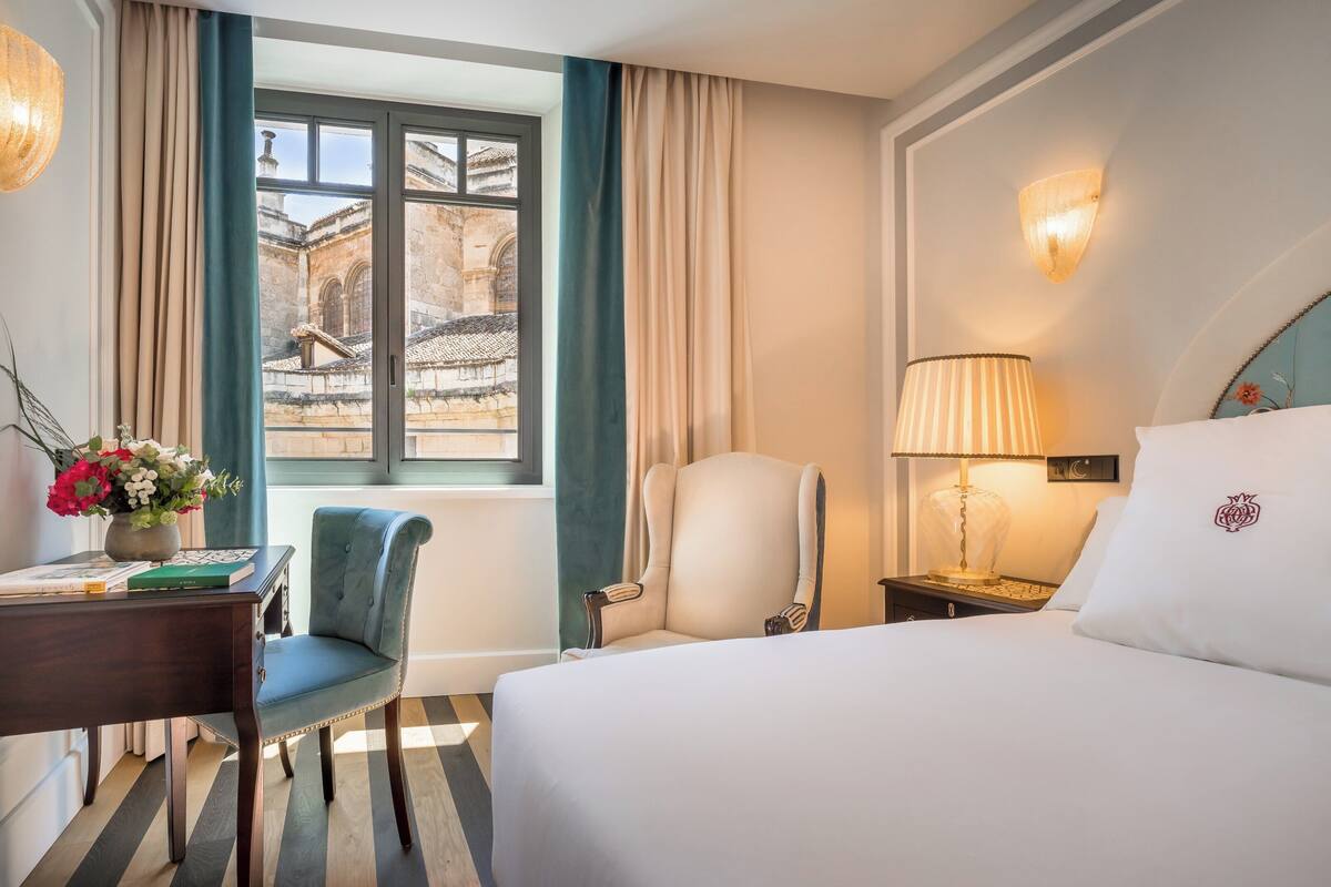 A hotel room with a bed, a desk, a chair, a lamp, and a view of a historic building through a window.