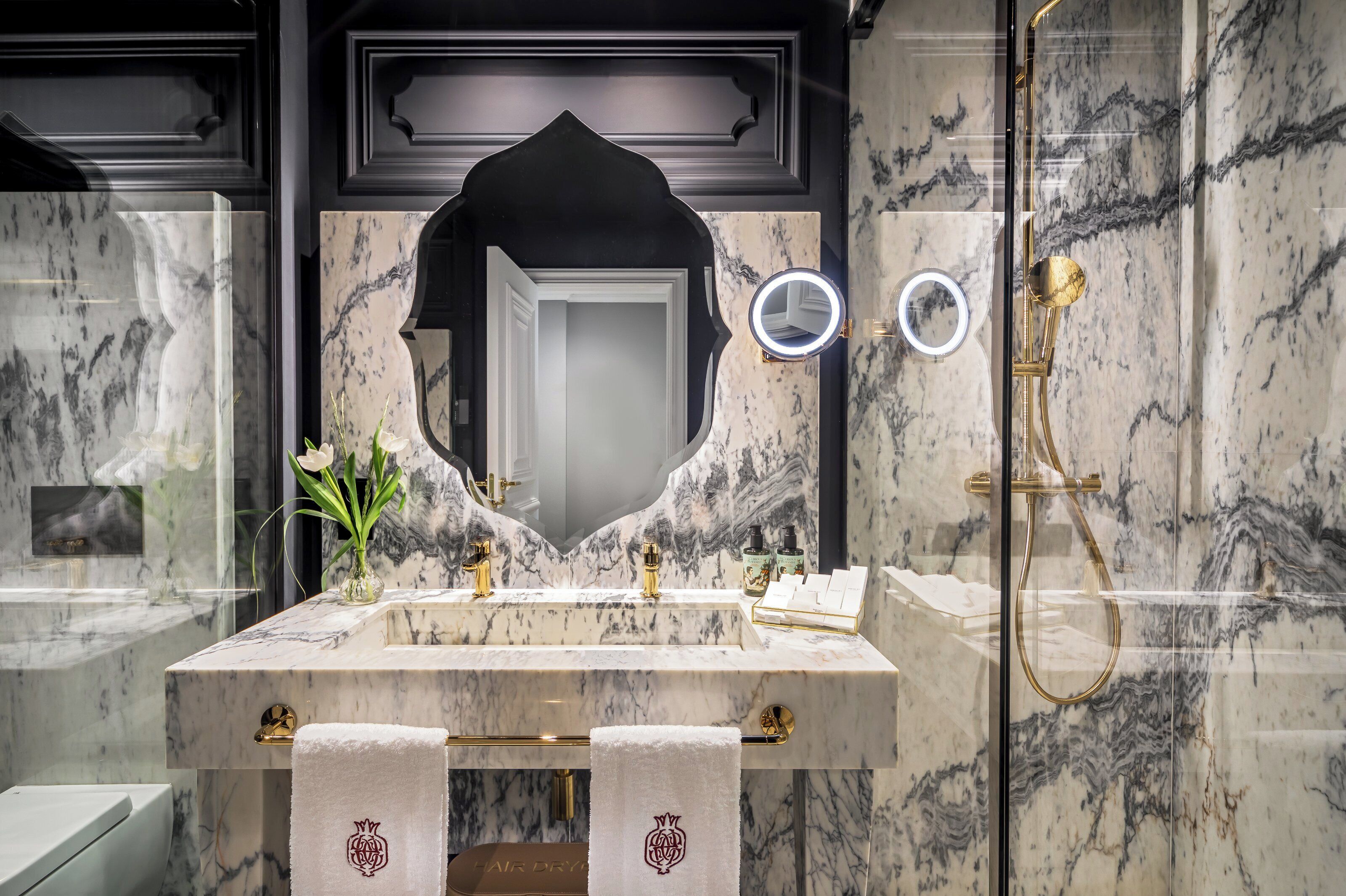 Une salle de bain en marbre avec un grand miroir, des accessoires dorés et une pomme de douche dorée.