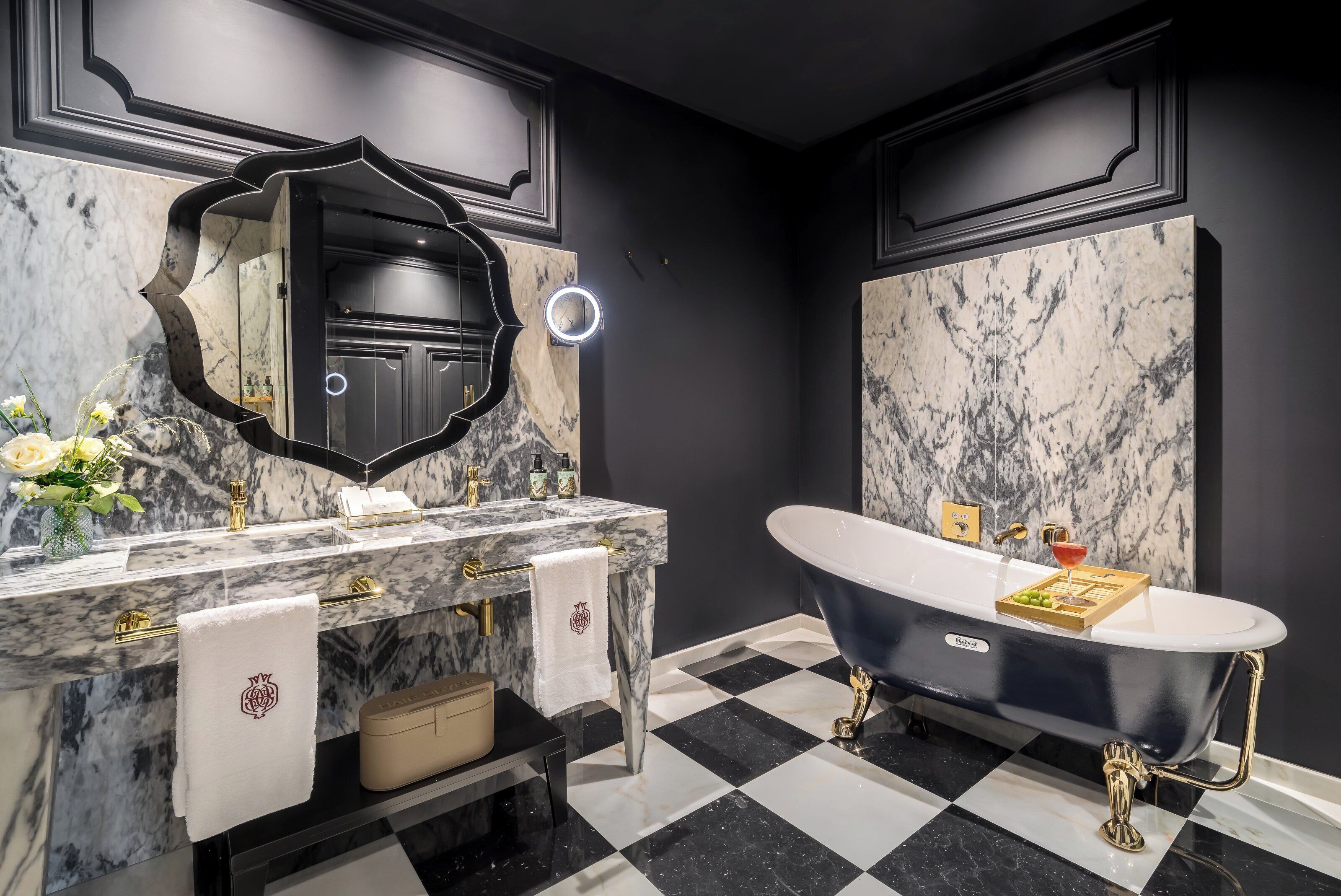 A bathroom with a black and white checkered floor, a freestanding bathtub, a marble vanity with gold fixtures, and a large mirror with a light.