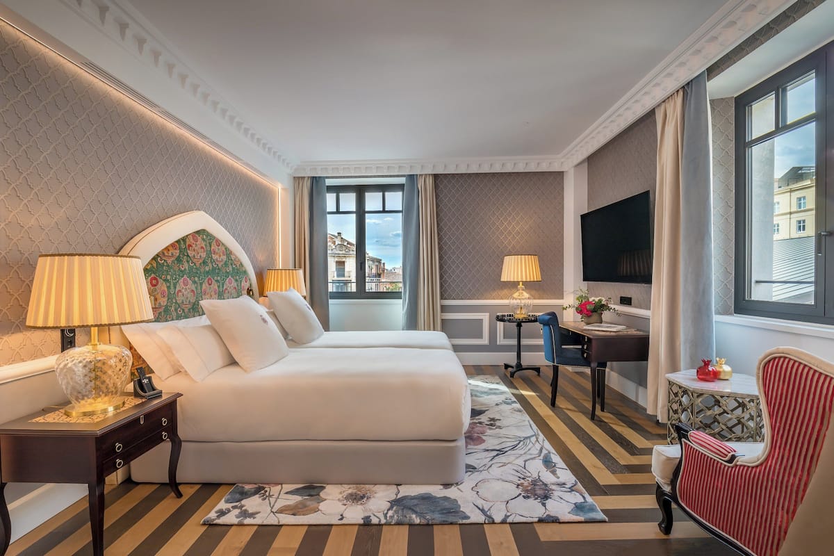 A hotel room with a bed, two bedside tables, a TV, a chair, and a window with a view.