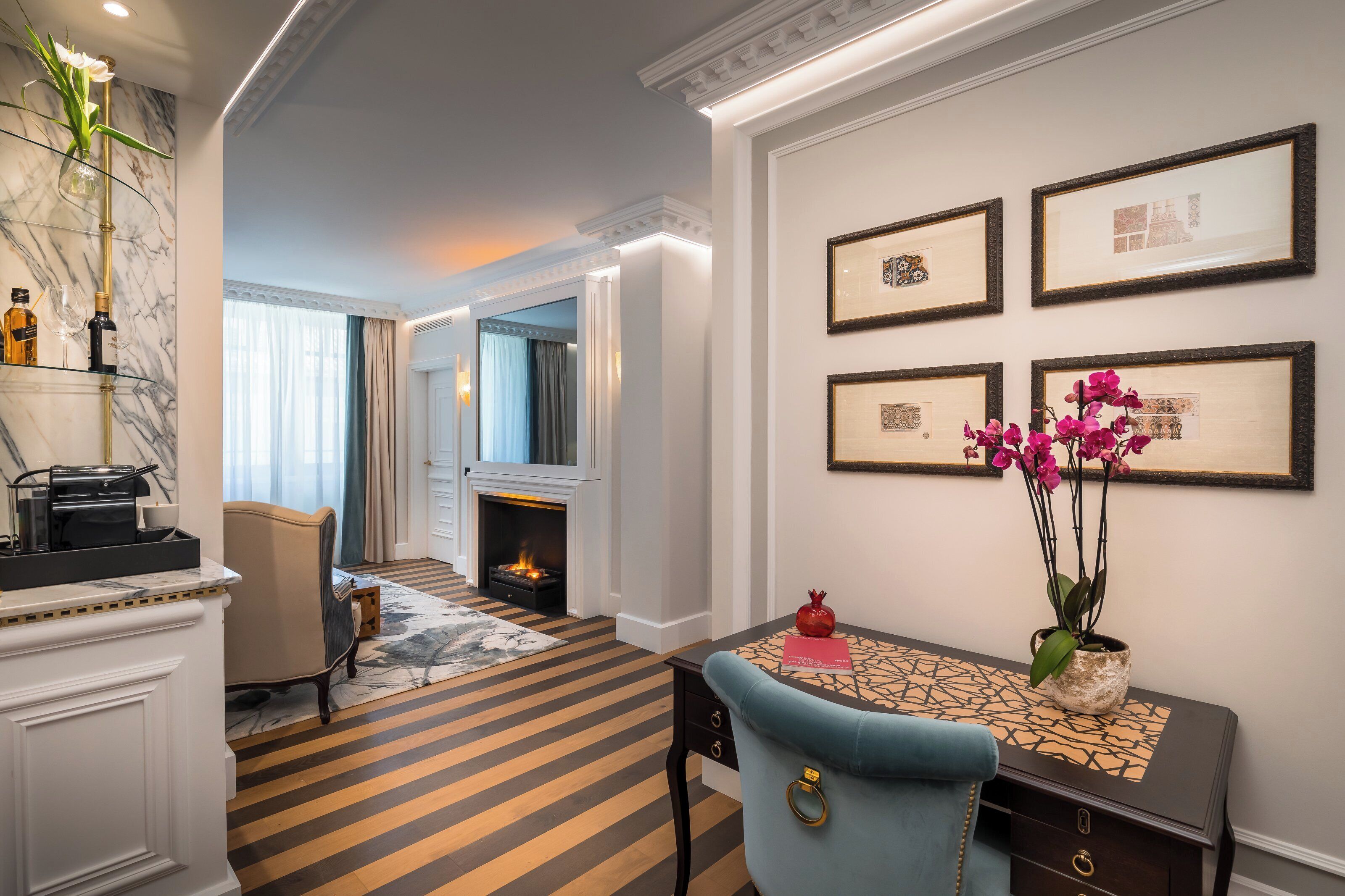 A hotel room with a fireplace, a dining area with a table and chairs, and a wall with framed pictures.